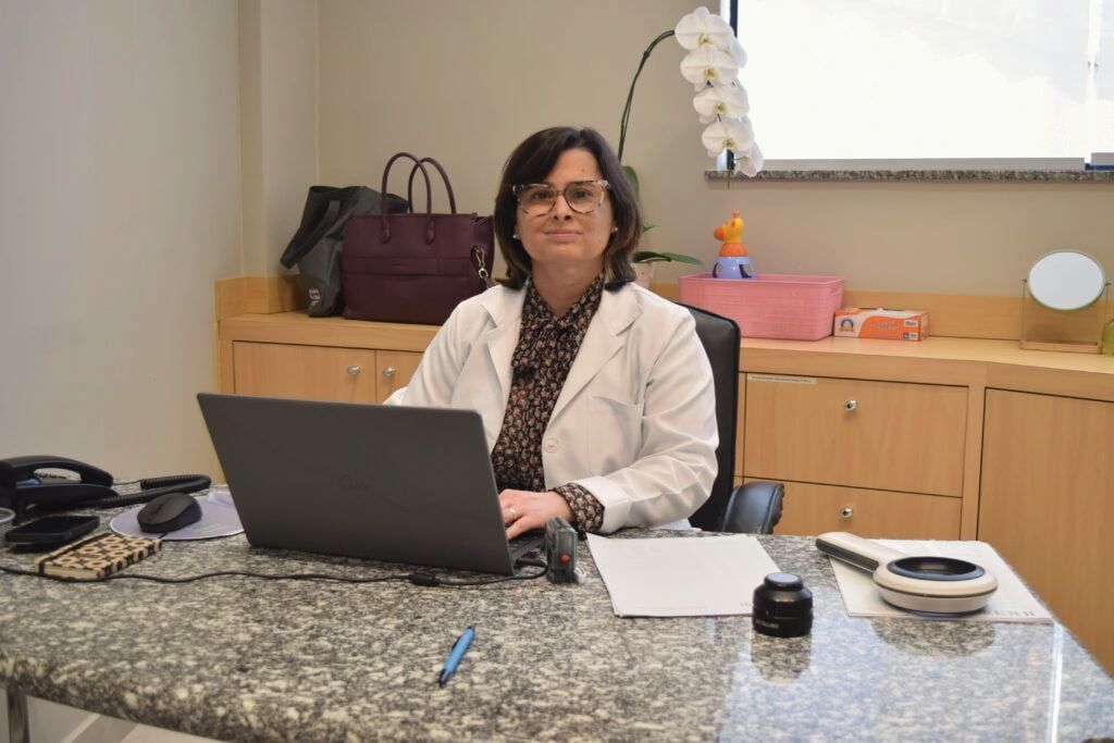 Dra. Daniela Gontijo à mesa do consultório, com orquídea e brinquedo infantil ao fundo